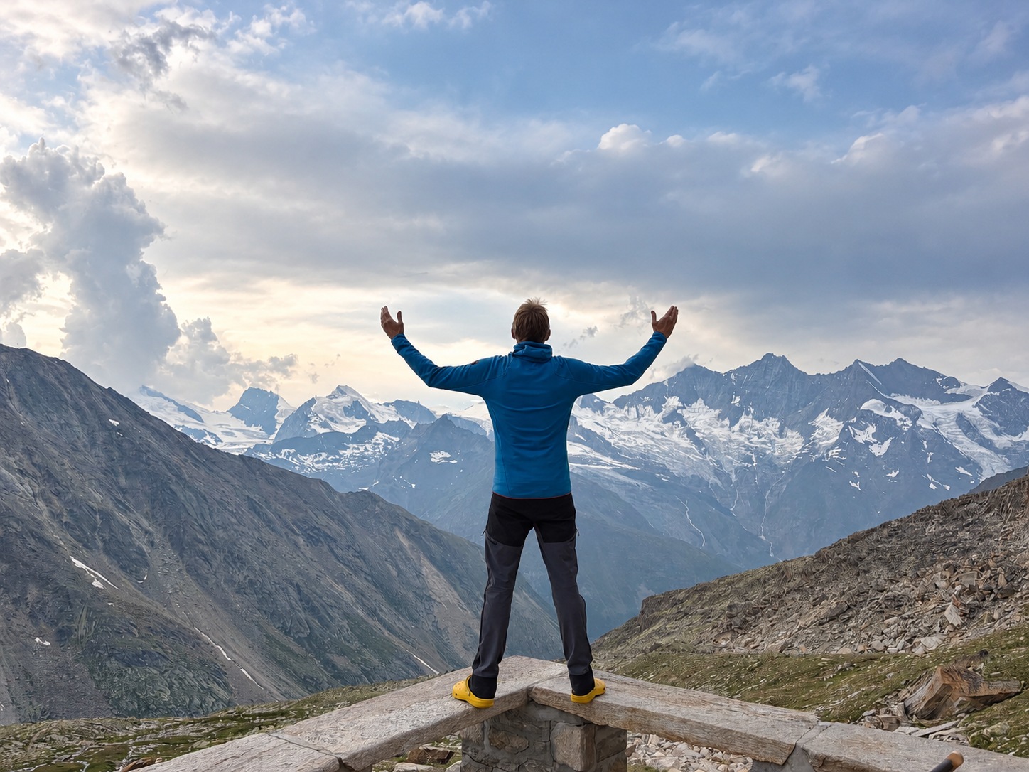Personne vue de dos, bras levés, face à un paysage de montagnes rocheuses et enneigées